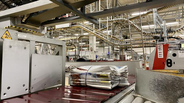 Die fertig gepackten Stapel werden automatisiert über Fließbänder transportiert. Im Hintergrund ist das komplexe Transportsystem zu sehen, das sich über den Köpfen der Mitarbeiter durch die Halle schlängelt | Foto: Kim Rileit