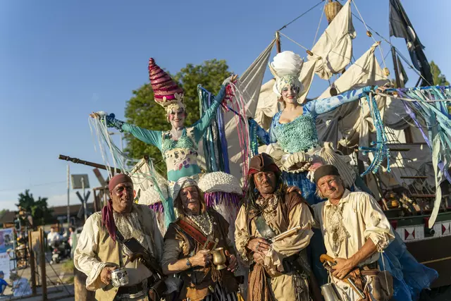 Vier der Kölner Piraten und die beiden Stelzenläuferinnen posieren vor einem nachgebauten Segelschiff. Die zwei Stelzenläuferinnen mit bunten Meerjungfrauenkostümen ragen über die Gruppe hinaus. Im Hintergrund: blauer Himmel und Festgelände. | Foto: ©projektart/Thomas Adorff