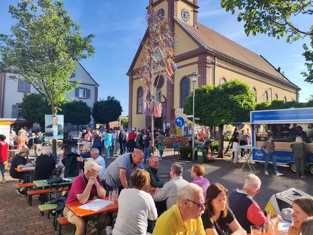 Das Bild zeigt Menschen, die auf Bierbänken sitzen. Im Hintergrund eine Kirche und ein Maibaum