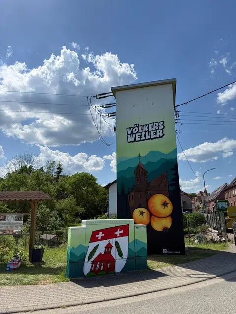 Alt-Text: Am Ortseingang von Völkersweiler steht ein neu gestalteter Umspannverteiler mit großflächigem Kunstmotiv. Zu sehen sind der Schriftzug „Völkersweiler“, die Dorfkirche, stilisierte Berge sowie drei goldgelbe Holzäpfel vor dunklem Hintergrund. Davor befindet sich ein kleinerer Verteilerkasten mit dem Ortswappen. Über der Anlage verlaufen Stromleitungen, im Hintergrund sind Häuser, Bäume und ein sonniger Himmel mit großen Wolken zu sehen.
