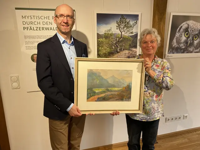 Frau Ulrike Bahl (rechts zu sehen) und der Museumsleiter des Museums unterm Trifels, Sven Gütermann, (links zu sehen) heben lächelnd gemeinsam das gestiftete Aquarell von Christoph Kröwerath in die Kamera. Das Aquarell zeigt im Hintergrund die drei Burgen Trifels, Aneobos und Scharfenberg, die rund um die Stadt Annweiler liegen. Die Burgenhügel sind in kühlen grün, gelb und blau Tönen gestaltet und als Umrisse erkennbar. Im Vordergrund ist in warmen Orange- und Brauntönen ein Weg mit Vegetation angelegt.