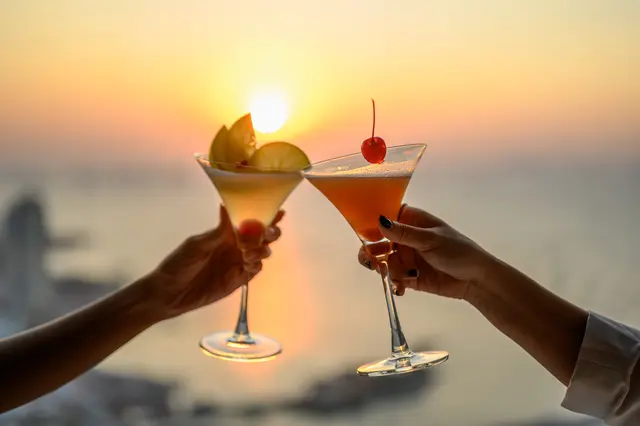das Bild zeige zwei Arme und Hände die Cocktailgläser halten und sich zuprosten