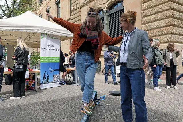 Eine junge Frau balanciert über eine Slackline, eine etwas ältere Frau stützt sie leicht am Arm. Im Hintergrund Infostand mit Roll-up "Werde Sportlehrer:in". Campus mit Besucher:innen.