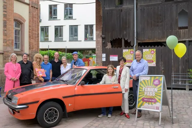 Der Heidelberger Ferienpass feiert 50. Geburtstag im Look der 70er. Baujahr 1976 ist auch der orangerote Opel Manta, um den sich als Geburtstagsgäste Oberbürgermeister Eckart Würzner (re.) und Bürgermeisterin Stefanie Jansen (2.v.r.) mit den „Zeitzeugen“ Aline Moser (li.), Alexander Espen-Zobeley (5.v.l.) und Toni L. (7.v.l.) gruppiert haben.  | Foto: Stadt Heidelberg/Philipp Rothe