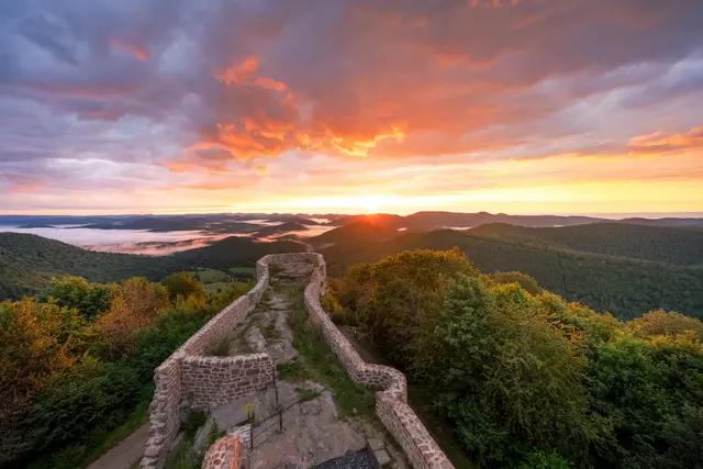 Das Foto zeigt den Blick Richtung Osten von einem oberen Betrachterstandpunkt aus. Dabei ist das unterhalb liegende Burgareal zu sehen. Dahinter erstreckt sich die Pfälzer Waldlandschaft mit kleinen Hügelketten und einem See zur rechten Seite, die in sanftem Morgenlicht beleuchtet werden. Das Morgenrot wird in den Wolken oberhalb der Burg reflektiert.