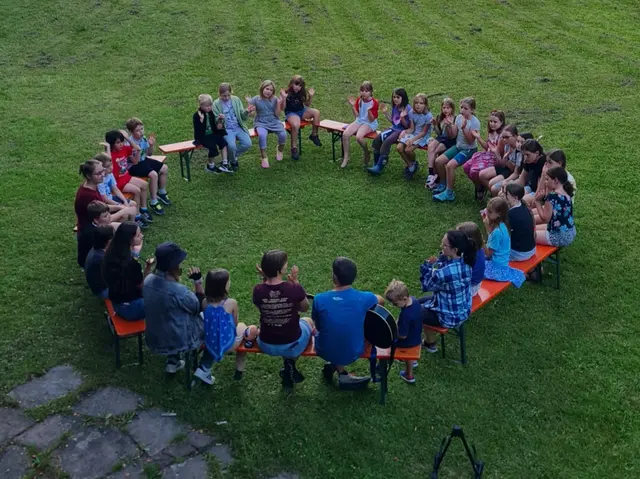 Eine Kindergruppe sitzt auf Bierbänken in einem Kreis, draußen auf einer Wiese.