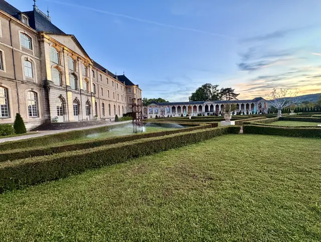 In den streng gezogenen, mit niedrigen Hecken gefassten Gartenkarrees der Abbaye des Prémontrés, die sich im sanften Abendlicht direkt an den Ufern der Mosel erstrecken, gehen die monumentale Symmetrie der meisterlich komponierten Klosterarchitektur und die leise Platzierung zeitgenössischer Kunstwerke eine tiefgründige Symbiose ein – ein in sich gekehrter Raum, in dem das jahrhundertealte Erbe klösterlicher Ordnung und die flüchtige Ästhetik der Moderne in einem stummen Dialog über die Dauerhaftigkeit der Zeit miteinander verschmelzen.
 | Foto: Daniel J. Basler