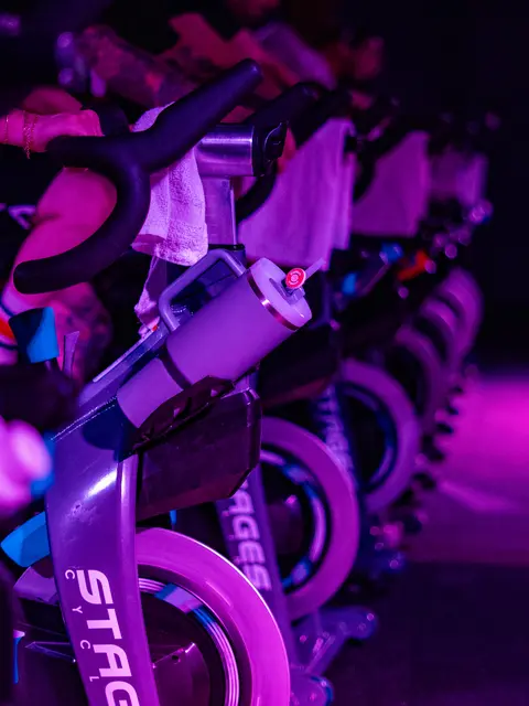 Nahaufnahme der Stages-Bikes bei Power Cycle Studio, in violettes Licht getaucht – bereit für die nächste Session. (Foto: Power Cycle Studio)  ALT-Text:  Reihe von Indoor-Cycling-Bikes der Marke Stages bei Power Cycle Studio in Kaiserslautern, mit Handtüchern und Trinkflaschen, in violett-pinkes Licht getaucht.
