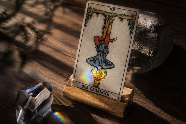 Die Tarotkarte 'Der Gehängte' in einem Holzständer auf einem Tisch, beleuchtet von Sonnenlicht und einem Regenbogenreflex eines Prismas.