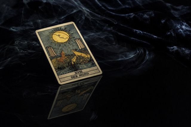 Die Tarotkarte 'Der Mond' spiegelt sich auf einer schwarzen Oberfläche, umgeben von feinem Rauch und dunklem Samt.