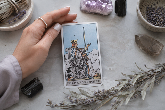 Eine Hand hält die Tarotkarte ‚Die Königin der Schwerter‘ auf einem hellen Untergrund, umgeben von getrocknetem Lavendel, Salbei und schützenden Heilsteinen.
