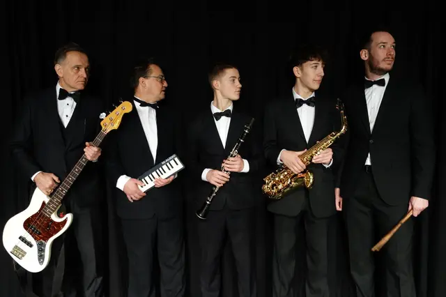 Fünf Musiker in schwarzen Anzügen, mit Fliege und Instrumenten.