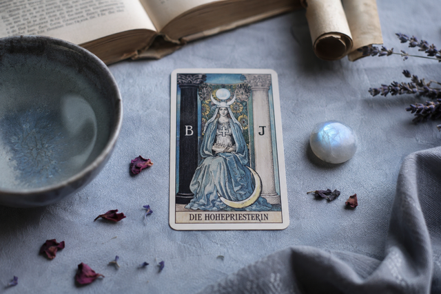 Die Tarot-Karte ‚Die Hohepriesterin‘ auf einer hellen Decke, umgeben von einem Mondstein, einer Schale Wasser, einem Lavendelzweig, trockenen Blütenblättern und einem alten Buch im sanften, kühlen Licht.