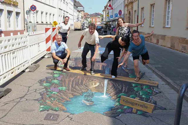Mit einem Fotopunkt mitten in der Baustelle wird die Königstraße in Landau zum beliebten Motiv. Martin Messemer, Leiter der Wirtschaftsförderung der Stadt Landau (v. l.), Lena Dürphold, Geschäftsführerin der Stadtholding Landau (h. l.), Manfred Stader, Straßenkünstler (2. v. l.), Nour Abboud, Vertreterin von Ralph Leibbrand, dem Ideengeber des Projekts (2. v. r.), Jana Schneider, Straßenkünstlerin (v. r.), und Nina Ziegler, Geschäftsführerin der StadtLeben (h. r.), werden selbst Teil des Kunstwerks. | Foto: Stadt Landau