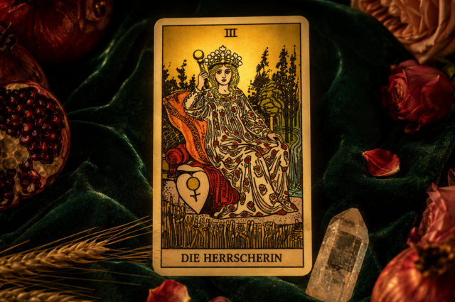 Die Tarot-Karte ‚Die Herrscherin‘ auf grünem Samt, umgeben von Granatäpfeln, Rosenblättern und Ähren im warmen Sonnenlicht.