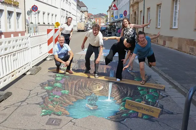 Mit einem Fotopunkt mitten in der Baustelle wird die Königstraße in Landau zum beliebten Motiv. Martin Messemer, Leiter der Wirtschaftsförderung der Stadt Landau (v. l.), Lena Dürphold, Geschäftsführerin der Stadtholding Landau (h. l.), Manfred Stader, Straßenkünstler (2. v. l.), Nour Abboud, Vertreterin von Ralph Leibbrand, dem Ideengeber des Projekts (2. v. r.), Jana Schneider, Straßenkünstlerin (v. r.), und Nina Ziegler, Geschäftsführerin der StadtLeben (h. r.), werden selbst Teil des Kunstwerks. | Foto: Stadt Landau