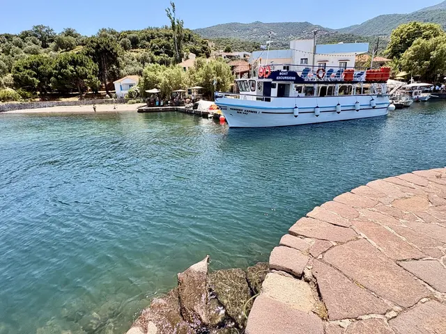 Ein Ausflugsboot ruht am Kai von Skala Sikamineas, Terrakottadächer klettern gemächlich den Hügel hinauf, Pinien werfen ihren Schatten über die Tavernentische, und ein paar Badende tummeln sich am Strand als gehöre die Zeit hier niemandem so recht. Gegenüber, außerhalb dieses Ausschnitts, wacht auf einem Felsplateau die Kapelle Panagia Gorgona über die Bucht — eine Madonna mit Fischleib, deren Herkunft kein Chronist kennt, deren Wirkung aber jeder Fischer des Ortes zu kennen glaubt. Stratis Myrivilis, geboren im nahen Sykaminia, verlieh ihr 1949 literarische Unsterblichkeit mit seinem Roman "Die Madonna mit dem Fischleib" (griech. „Η Παναγιά η Γοργόνα") — einer Geschichte über Heimkehr, Meer und den schmalen Grat zwischen Aberglaube und Glauben. Wer heute am Kai steht, sieht nur ein Boot, ein paar Häuser, das Wasser: die Kapelle bleibt unsichtbar und ist doch, wie jede gute Legende, längst überall im Bild.
 | Foto: Daniel j. Basler