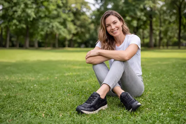 Virtuelle Caprice-Markenbotschafterin ANNA sitzt mit schwarzen Schuhen auf einer Wiese im Park.