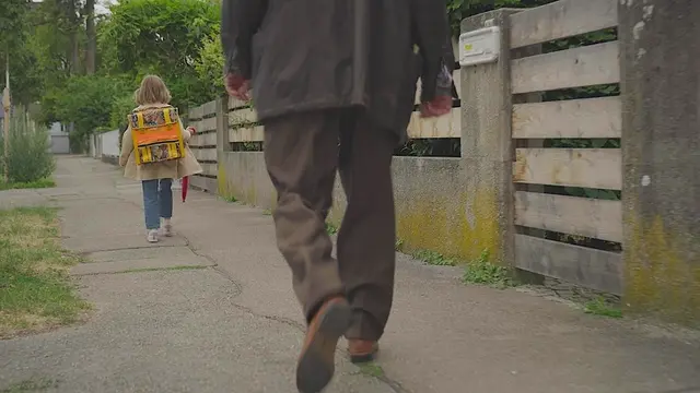 "XY gelöst - Der letzte Schulweg": Ein Schulkind mit auffälligem gelbem Schulranzen geht auf einem Gehweg durch ein Wohngebiet. Das Kind hält einen geschlossenen roten Regenschirm in der Hand. Im Vordergrund folgt in einiger Entfernung eine erwachsene Person auf demselben Weg. Rechts verläuft ein Zaun entlang der Grundstücke, links stehen Bäume auf Grünflächen.