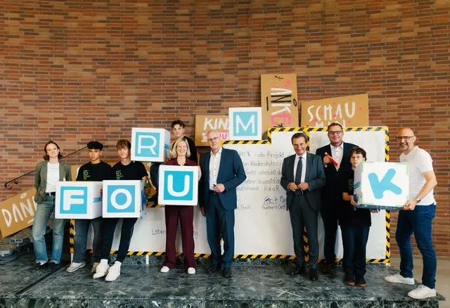 Gruppenfoto zum Baustellenfest des Forum K in Karlsruhe: Jugendliche und Erwachsene präsentieren gemeinsam Buchstabenwürfel, die „FORUM K“ ergeben, vor einer Backsteinwand mit kreativen Plakaten zum Thema Kinderrechte.