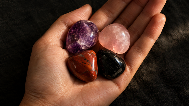 Eine Handfläche hält vier glatt geschliffene Edelstein-Trommelsteine – Amethyst, Rosenquarz, roter Jaspis und schwarzer Turmalin – vor einem dunklen Leinenhintergrund