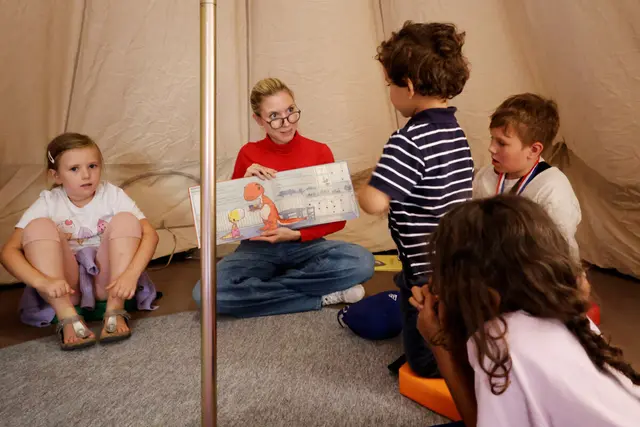 Eine Frau in der Mitte eines Zelts zeigt eine Doppelseite aus einem Kinderbuch, umringt von mehreren Kindern.