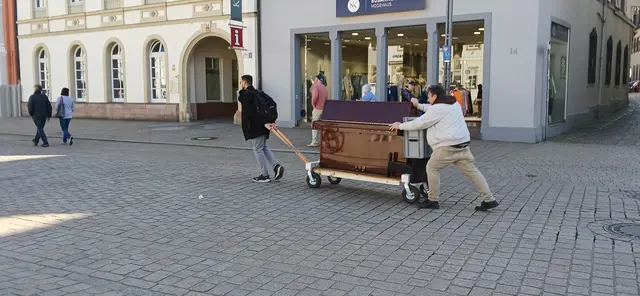 Auf der Hauptstraße Maximilianstraße in der Stadt Speyer wurden Passanten Zeugen einer ungewöhnlichen „Konzerttour ohne Plakat“.
Zwei entschlossene Männer legten einen improvisierten Eilmarsch hin und transportierten ein Klavier über das Kopfsteinpflaster mit so viel Enthusiasmus, als würden sie sich zu ihrem eigenen Zugaben-Auftritt verspäten.
Der eine zieht das Instrument nach vorn, der andere schiebt von hinten — und das alles ohne einen einzigen Ton, aber mit ausgezeichnetem Rhythmus.
Die Einheimischen fragten sich vermutlich:
„Ist das eine Hauslieferung von Musik oder eine neue Form von Straßen-Fitness?“
 | Foto: Liubov Arnold